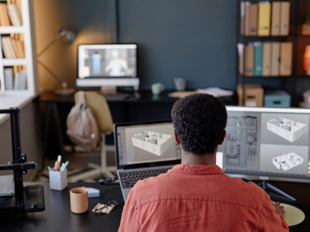 Person sitting at a desk working on architectural and 3D landscape designs displayed on dual monitors, with another computer and shelves in the background.