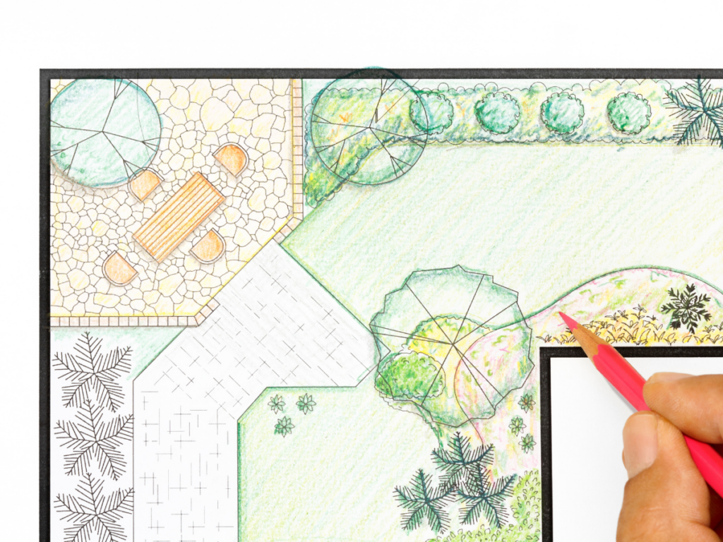 A hand uses a red pencil to draw on a landscape design plan featuring trees, shrubs, a walkway, and a patio with a table and chairs, providing the initial sketch before creating detailed 3D landscape designs.