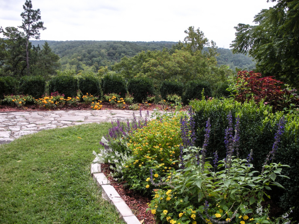 A stone path showcases thoughtful walkway design as it curves beside flower beds with colorful blooms and greenery, overlooking a forested landscape and distant hills under a cloudy sky.