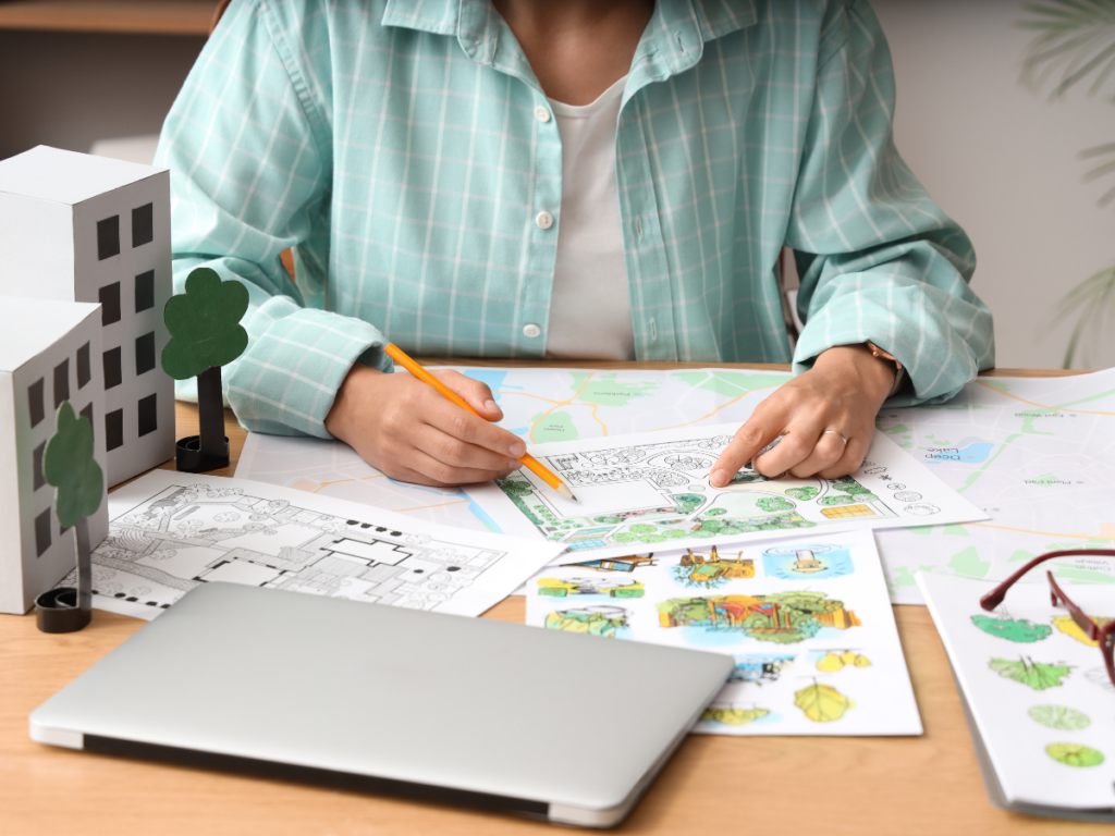 Person working at a desk with architectural models, floor plans, colored drawings, and a laptop, pointing at landscape design sketches and discussing 3D landscape design concepts.
