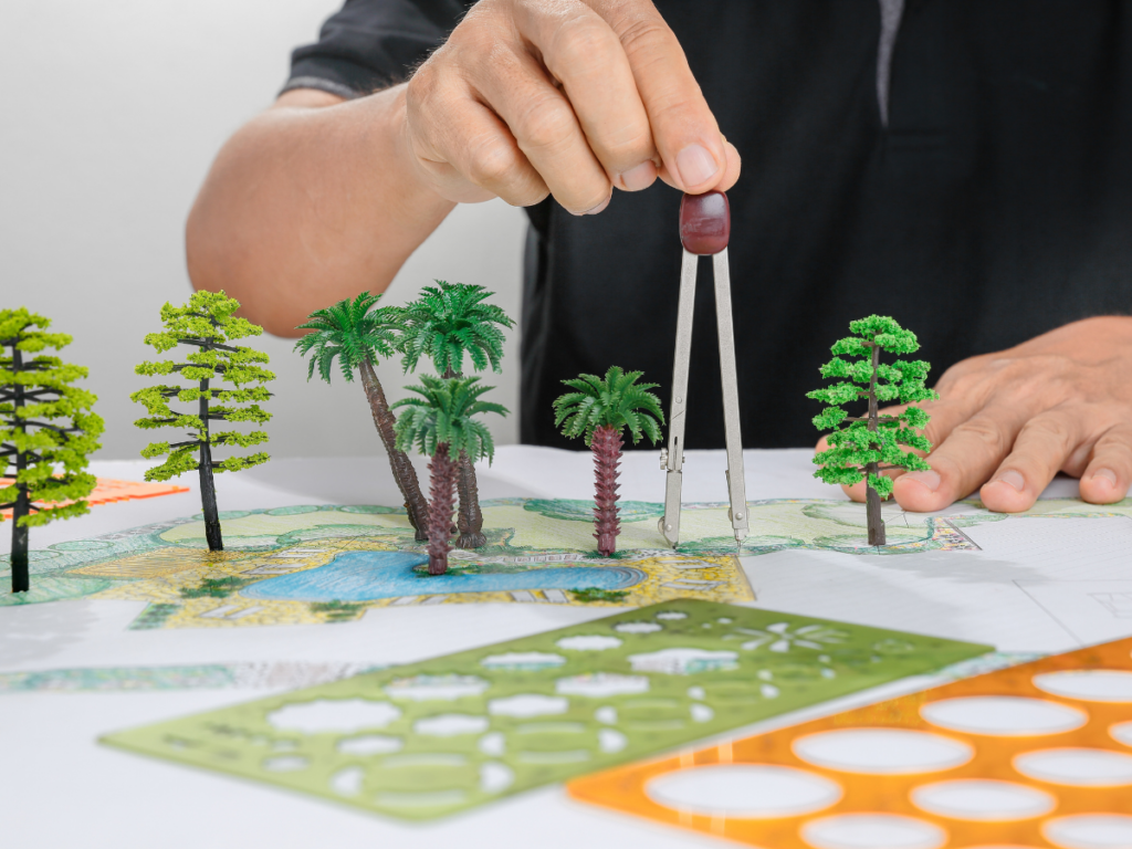 A person arranges miniature trees on a landscape design blueprint, using a compass and stencils to plan the layout, bringing their vision to life with principles of 3D landscape design.