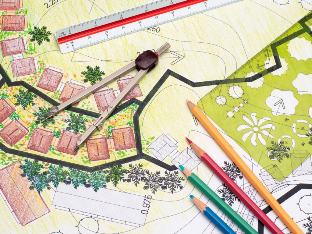 Colored pencils, a ruler, and a compass rest on a detailed 3D landscape design plan with buildings, paths, and vegetation sketched and colored in various shades.