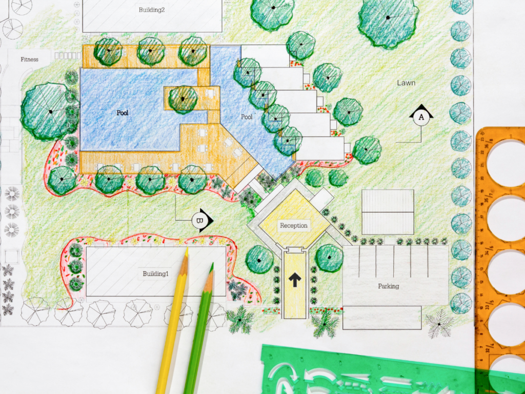 A colored architectural site plan illustrates a building complex with pools, a reception area, paths, lawns, trees, and parking. Drawing tools and colored pencils are placed on top, highlighting the detailed approach taken in creating this 3D landscape design.