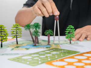 A person arranges miniature trees on a landscape design blueprint, using a compass and stencils to plan the layout, bringing their vision to life with principles of 3D landscape design.